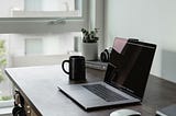 A home office setup. A desk sits next to a window. It has a laptop with a mouse, a coffee cup, a book upon which sits a plant and a camera, and other knickknacks. A pair of headphones hangs from one of the desk legs.