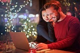 Interracial couple at a restaurant with laptop opened. The woman leans on man’s shoulder. Fairy lights in the distance.