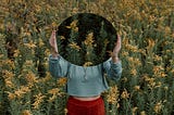A woman standing in a field of wildflowers, holding up a mirror to cover her face.