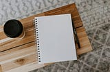 A blank notebook, a pen, and a cup of coffee sit on a wooden desk.