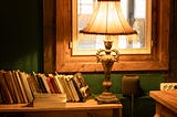 A bookshelf with a lighted lamp on it, with a window in the background.
