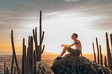 A hiker perches atop a rocky climb at sunrise. He’s alone, but in the company of tall, thin spires of cacti rising up around him. A city sprawls in the distance beneath the soft, golden hued sky.