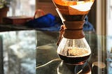 A Chemex coffee maker brewing coffee, with steam rising from the top. The warm sunlight illuminates the scene, casting reflections on the glass table. A blurred background with greenery and a framed picture add a cozy atmosphere.