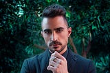 Sleek dressed male with side cuts hairstyle and trimmed beard, wearing blazer, watch, looking into camera determined.