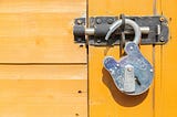 A heavy-duty silver padlock hanging completely open and unlatched from a dark metal slide bolt lock on a bright, sunlit yellow wooden door. The open lock sits on the right side of the horizontal frame, symbolizing freedom, broken silence, and midlife liberation