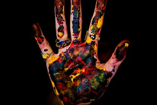 A hand, held up, palm facing, covered in paint, on a black background.