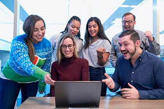 Six people of different ages, sexes, and races are smiling excitedly looking at a laptop screen