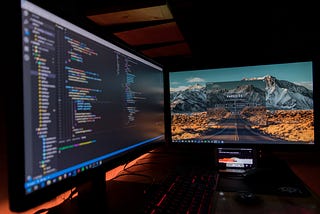 A photo of a monitor with some code on it and a second monitor with a website and a phone playing a song.