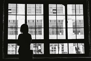 A silhouette of a person standing by a large window, looking out at a building with multiple windows on the opposite side. The scene is captured in black and white, highlighting the contrast between the dark interior and the bright exterior. The person appears contemplative, adding a sense of introspection and solitude to the image.