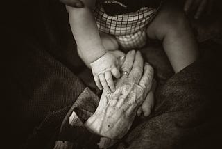 black and white close up of an elderly hand, ring on the wedding finger. There is the lower half of a very small child with their hand on top of the elder.