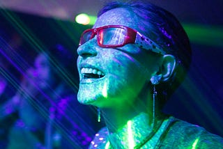 This image depicts a person at what seems to be a party or event where UV lighting is in use, as indicated by the vibrant greenish glow on their face and clothing. The person is wearing red-framed glasses and earrings, and appears to be having a joyful moment, with their mouth open as if they are laughing or cheering. The background is blurred, but lights and possibly other people can be seen, suggesting a lively atmosphere. The visible laser lights add to the festive ambiance of the setting.
