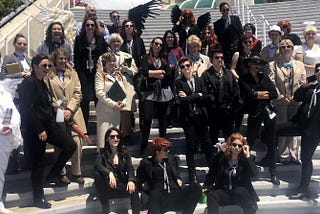 A photo of a large group of Good Omens cosplayers. They are mostly Crowley and Aziraphale cosplayers with a few other characters as well. No two of them look the same, despite the fact that most of them are cosplaying the same two characters.