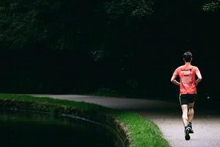 A man in a red shirt and black shorts running on a paved, winding path alongside a body of water