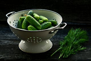 A metal colander on a black surface with a black background. Inside, a pile of small, bright green cucumbers, next to it, a dark green bunch of fennel.
