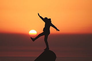 sillhouette of someone standing on one foot on a rock in front of the sunset