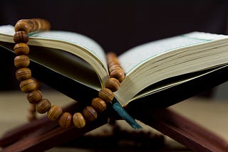 Image of white pages of an open Bible sitting on a wooden stand with wooden rosary beads laying on top of it. The image was chosen by Spiritual Lifestyle Writer, The Christine McDonald, to highlight how to figure out your spiritual life purpose and where you get the strength to follow it.