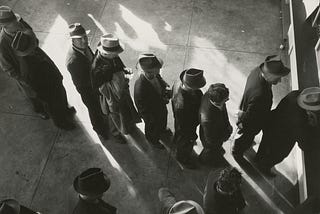 Black-and-white photo showing several people standing in a line for work, viewed from above. They wear hats and coats and appear to be waiting near a doorway.