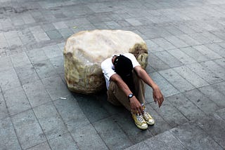 A Black man in casual clothes and sneakers, sits alone on the ground, with his back against a boulder. His head rests between his knees. He is tired.