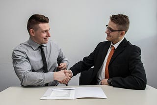 Two men in suits shake hands in an interview-style room.