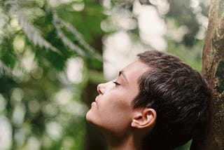 Person leaning their head back against a tree with eyes closed. They are feeling the sun on their face and look relaxed and at ease in the forest around them.