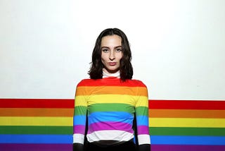 A young woman wearing a rainbow shirt that almost blends into the rainbow painting on the wall.