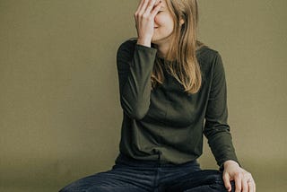 A woman sitting criss-cross applesauce with her right palm covering her face in a mixture of embarrassment and laughter.