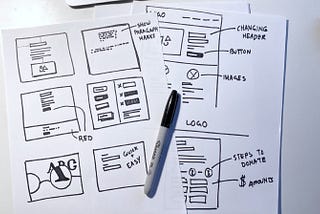 Rough drawings of computer screens on white paper sitting on a desk. A black pen rests on top of them.