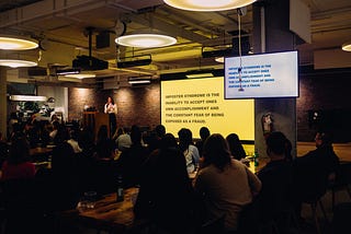 A woman standing next to a large projection of a slide. The slide contains a statement about imposter syndrome. A large audience is also in the foreground.