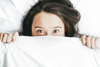 A terrified woman, with a white bedsheet pulled over her eyes. You can see her brunette hair, her face upto her eyes, and two hands clutching the bedsheet.