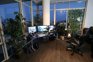 Picture of an office, desk in front of window, three computer monitors on.