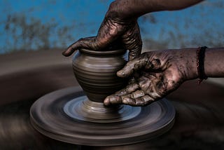 Close up of a vase being thrown on a potter’s wheel against a turquoise wall.