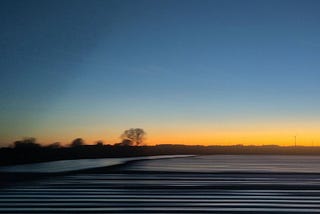 Sunset seen from a moving train. In the foreground horizontal marks across a ploughed field, at the horizon a row of about 5 trees, slightly out of focus, one larger than the rest. The sky is a gradient from orange at the bottom to dusty deep blue at the top