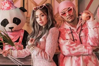 Duas pessoas de rosa. Com um panda de de chapéu rosa. Os cantores Kimbeley Chan e o rapper namewee. Ela com um estilo todo modelo com óculos na cabeça; ele com óculos rosa e toca e moletom, segurando uma maçã.