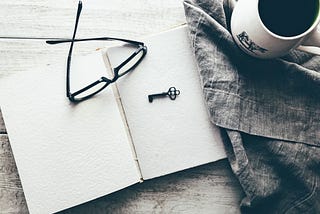 A black and white image of an open book with glasses and a key on it, and next to it a cloth and a cup of coffee.