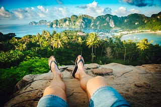 A man wearing flip flops, and lying on a rock in a tropical setting.