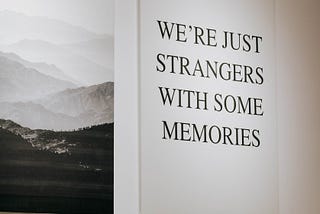 Large canvas on a wall in a gallery with nothing but the following text on it in all caps: We’re just strangers with some memories.