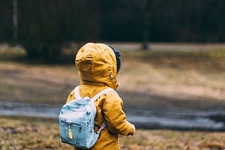 A child walking down a hill in early spring, with an orange coat and hood, and a light blue backpack.