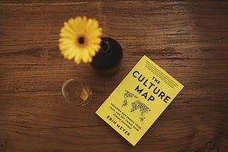 A yellow daisy in vase next to white wine glass and yellow book titled, “The Culture Map” by Erin Meyer on a brown table.