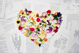 Multi-colored flower petals are in the shape of a heart. The heart is on top of book pages with black and white text.