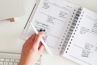Hand writing in a planner next to a laptop and keyboard on a white desk.