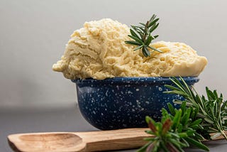 a bowl of vanilla ice cream with a sprig of rosemary and a wooden spoon nearby.