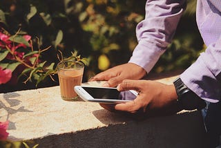 Man sitting on a ledge with a coffee next to him. He is scrolling on his Apple iPhone, probably for way too long.