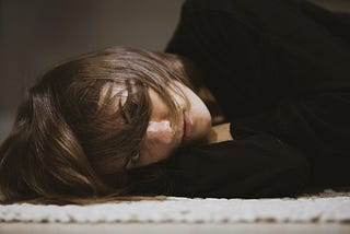 A woman with dark hair and wearing a black jumper lying on the floor, her hair across her face.