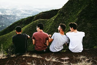 Four males sitting side by side in nature — laughing