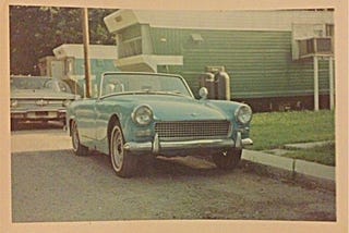 Old green Austin Healey convertible parked in front of a mobile home