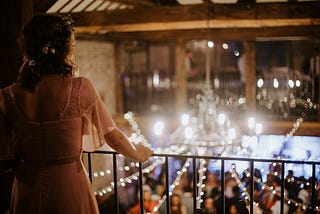 Photo of a woman in a pink dress looking over a balcony railing past fairy lights and a chandelier into a formal event.