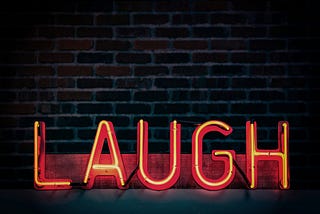 an illuminated red and yellow neon sign of the word LAUGH against a brick wall background