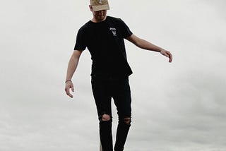 Man in black wearing a cap, balancing on a beam, with ocean in the background