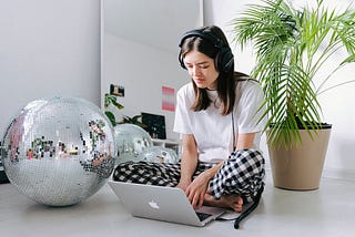 Woman in white shirt and pajamas using MacBook next to a disco ball and plant