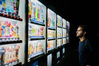 An image of a Human looking at a six panel wide vending machine and looking confused about what choice to make.